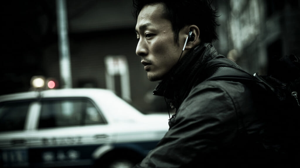 自転車で片耳イヤホンを装着しながら走行する日本人男性と背後を走る車の様子