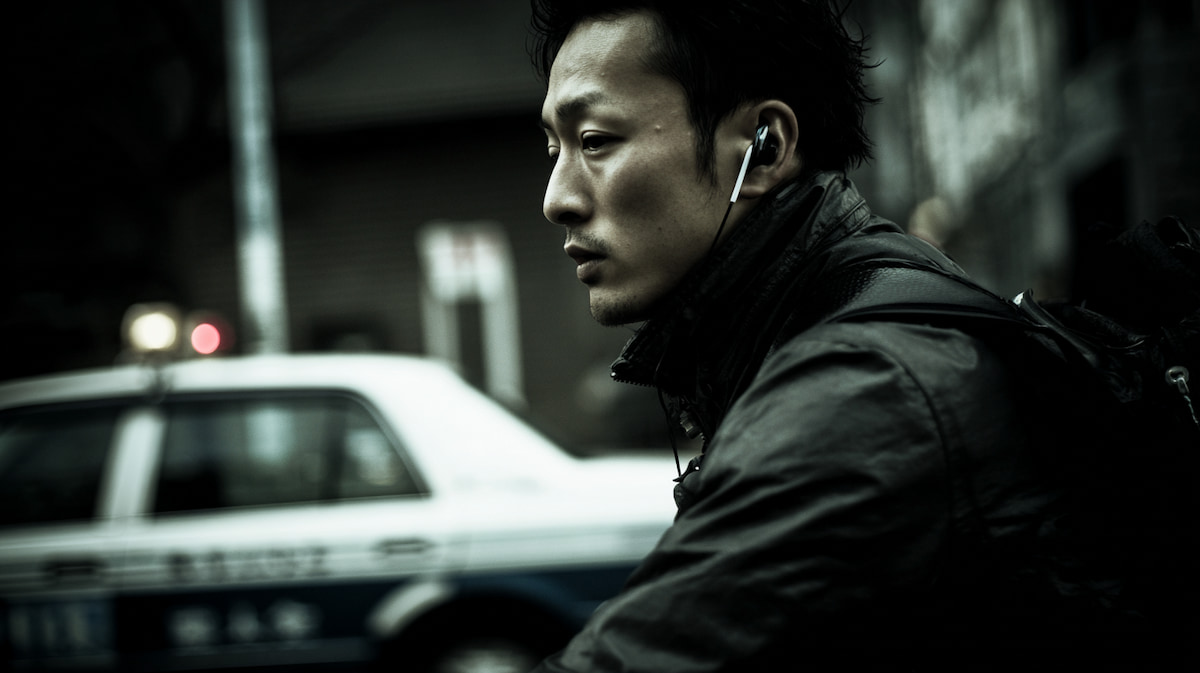 自転車で片耳イヤホンを装着しながら走行する日本人男性と背後を走る車の様子